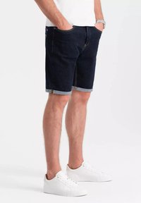 Shorts de mezclilla en azul oscuro, con un corte recto, puños enrollados y bolsillos laterales. Combinados con zapatillas blancas.