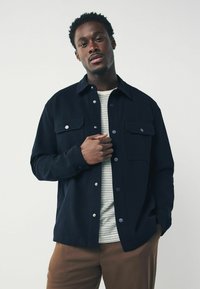 Chaqueta de botón azul marino con dos bolsillos en el pecho, botones a presión plateados y una textura suave, llevada sobre una camisa a rayas y pantalones marrones.