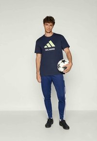 T-shirt navy con testo verde "REAL MADRID" e logo a tre strisce, abbinato a pantaloni blu con bande laterali bianche, che tiene in mano un pallone da calcio in bianco e nero.