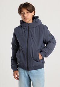 Teenage boy wearing a navy blue hooded jacket and light blue jeans, standing with one hand in pocket against a plain background.