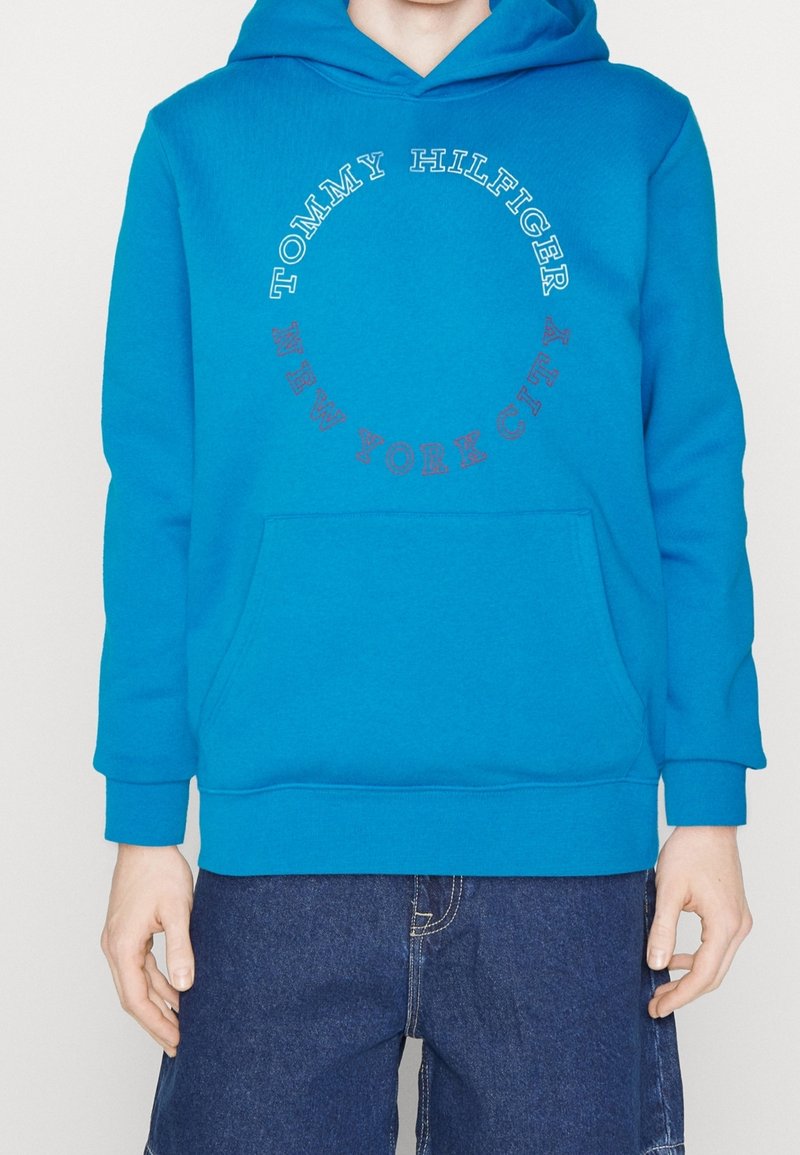 Personne portant un sweat à capuche Tommy Hilfiger bleu vif avec le texte "TOMMY HILFIGER" et "NEW YORK CITY" disposé en cercle, et un short en jean bleu foncé.