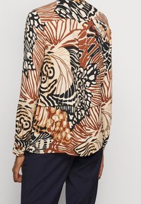 Blusa stampata in tonalità beige, marrone e nera. Caratterizzata da un motivo floreale, maniche lunghe e una vestibilità rilassata con yoke posteriore e dettaglio con bottoni.