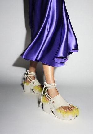 Scarpe bianche con punta aperta e dettagli in morbido giallo, tacchi a blocco alti e lacci incrociati. Completate da una gonna brillante in satin viola.