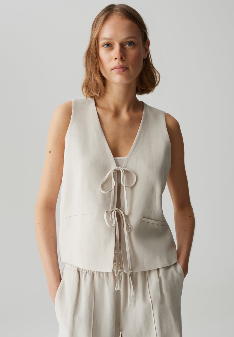 Gilet senza maniche beige chiaro con scollo a V, chiusura frontale con cordoncino e due tasche frontali. Tessuto morbido e testurizzato con vestibilità rilassata.