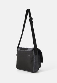 Bandolera con un diseño de tela negro y gris, que cuenta con un cierre de solapa, una correa ajustable y un bolsillo frontal con cremallera para almacenamiento.