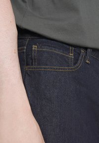 Ciemnoniebieskie dżinsy z kontrastującym żółtym przeszyciem, z kieszenią z przodu i prostym krojem; w połączeniu z szarym T-shirtem.