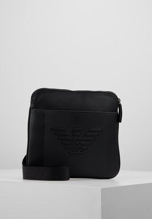 Sac bandoulière rectangulaire noir avec logo embossé sur le devant, fermeture éclair et sangle d'épaule réglable, présenté sur un socle blanc.