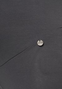 Tecido cinza escuro com uma textura suave, apresentando um detalhe metálico circular prateado com texto gravado. O design inclui costuras anguladas.