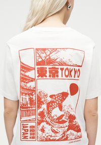 Osoba o blond włosach w białej koszulce z czerwonym nadrukiem przedstawiającym karpia koi, Górę Fuji, pagodę oraz tekst „Tokyo” i „Japan” na plecach.