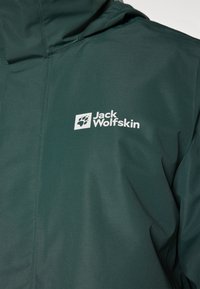 Donkergroene Jack Wolfskin-jas met een soepele textuur, capuchon en een opvallend wit logo met een pootafdruk op de linkerborst.