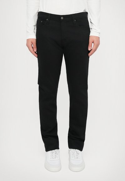 Pantaloni in cotone neri con vestibilità dritta, tasche frontali e chiusura con bottone, abbinati a sneaker bianche. Design e texture semplici.