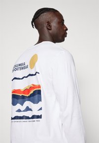 Pika pikkade varrukatega valge T-särk, millel on värvikas mägigraafika; esindatud sinised, oranžid ja kollased jooned ning seljal "Columbia Sportswear" tekst.