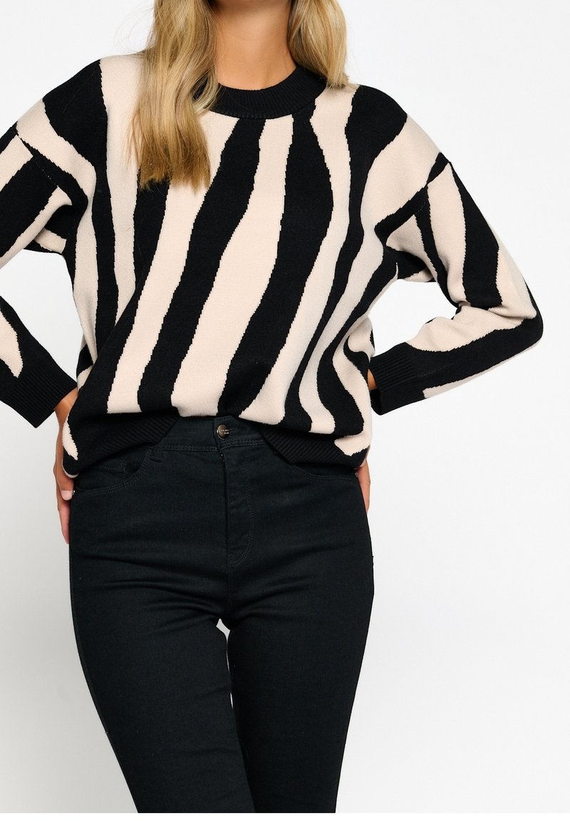 Suéter a rayas negro y beige con textura de punto y cuello acanalado, combinado con jeans ajustados negros. Corte relajado y mangas largas.