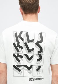 T-shirt de algodão branco com uma estampagem gráfica nas costas, apresentando formas e linhas geométricas pretas, marca "KARL LAGERFELD JEANS."