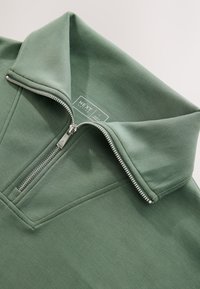 Green zip-up pullover with a high collar and silver zipper. Made from smooth, soft fabric with a subtle texture. Brand label visible.