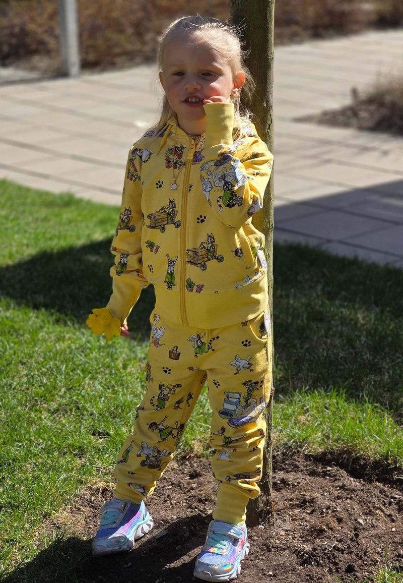 Gul zip-up huvtröja och matchande byxor med tecknade djur och objekt, kombinerat med iridescerande sneakers, stående vid ett träd.