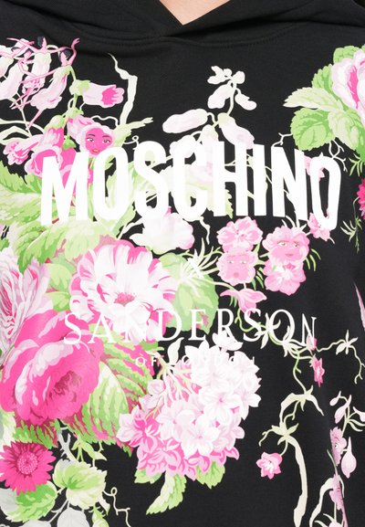 Melna sporta krekliņa priekšpuse rotāta ar spilgti rozā un baltu ziedu rakstiem. Priekšpusē ir drosmīgs balts teksts: "MOSCHINO SANDERSON of LONDON."