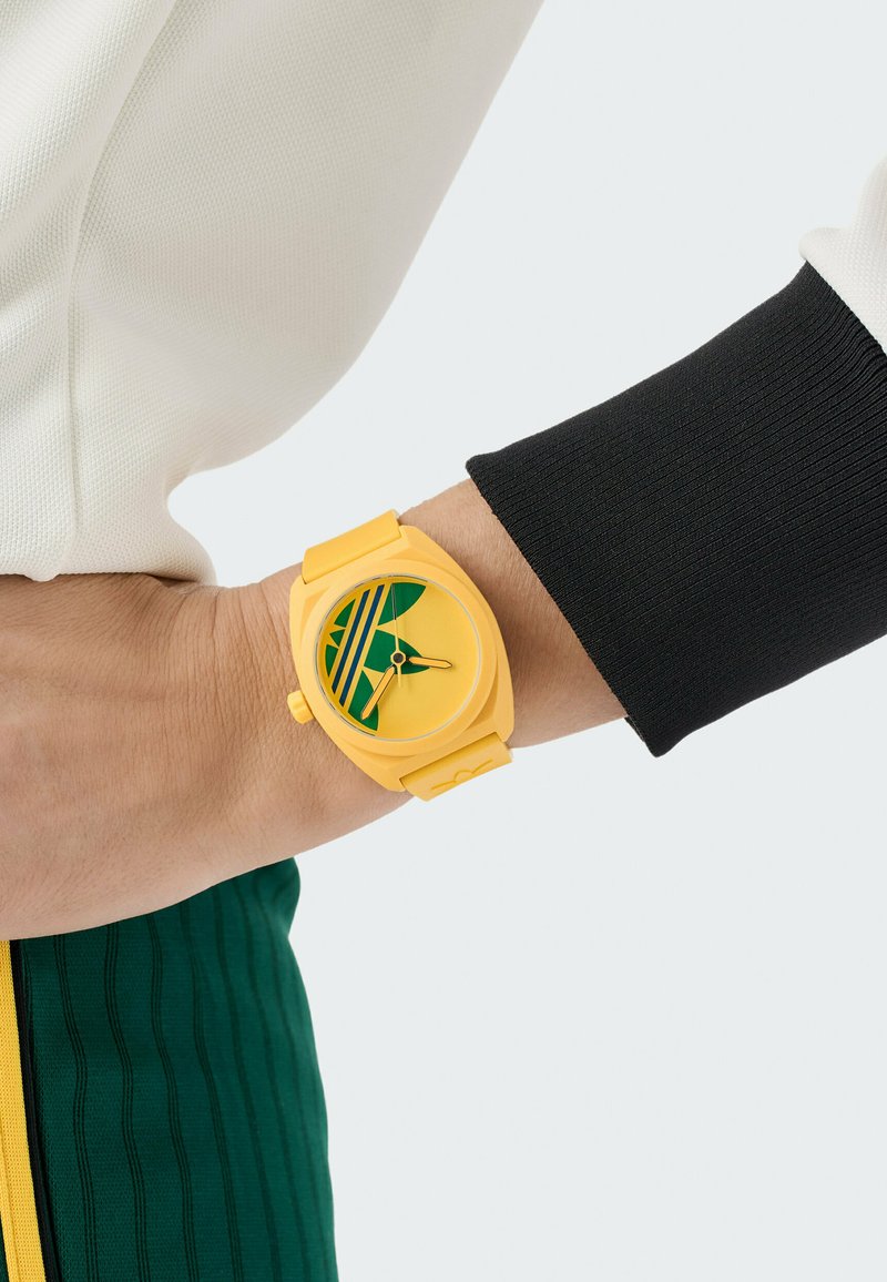 Gele horloge met een ronde wijzerplaat, voorzien van groene accenten en een gestreept ontwerp. Het materiaal lijkt rubberachtig met een gladde textuur.