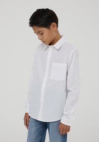 Camicia bianca a maniche lunghe realizzata in tessuto leggero, con chiusura a bottoni frontale, colletto standard e una singola tasca sul petto.