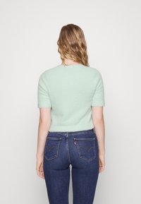 Mátově zelený krátký chlupatý svetr s kulatým výstřihem. Nošený s tmavě modrými džínami s kapsami na zadní straně a štítkem Levi's.