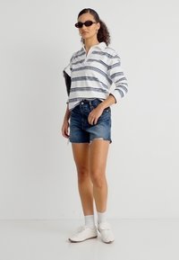 Γυναίκα που φοράει μακρυμάνικη ριγέ πόλο, κοντά τζιν σορτς, λευκά αθλητικά παπούτσια, λευκές κάλτσες, γυαλιά ηλίου και κρατάει μια μαύρη τσάντα ώμου.