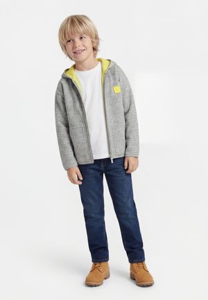 Smilende blond dreng iført en grå lynlåshættetrøje med gul foring, hvid t-shirt, blå jeans og beige snørestøvler, stående med afslappet kropsholdning.