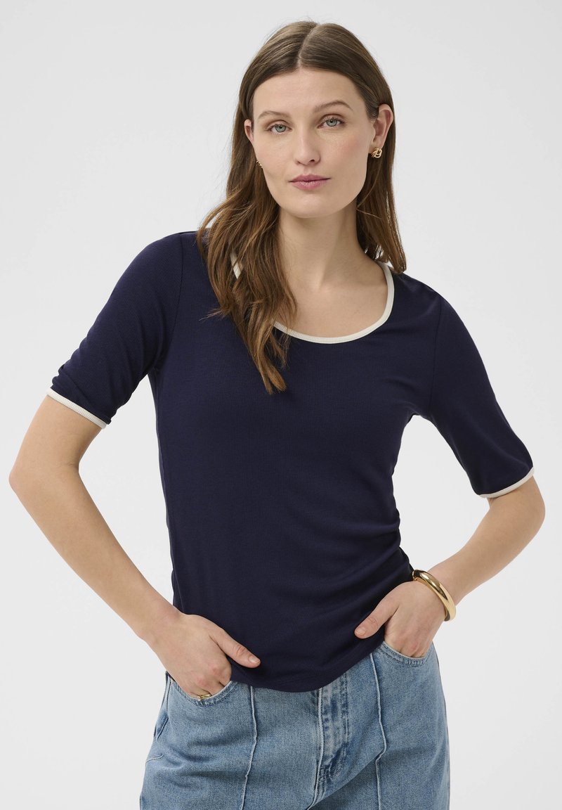 Mujer con cabello castaño que lleva una blusa azul marino con ribete blanco y jeans azul claro, de pie con las manos en los bolsillos frente a un fondo liso.