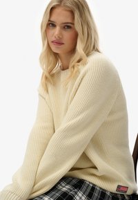 Donna bionda che indossa un maglione lavorato a maglia color crema e una gonna scozzese bianco e nera, seduta con una leggera inclinazione e un'espressione neutra.
