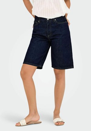 Bermuda shorts en denim bleu foncé avec une coupe droite, un design à cinq poches et des détails de couture dorée. Associés à un top blanc et des sandales.