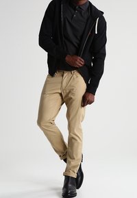 Svart zip-up hoodie över en mörk pikétröja, khakibyxor och svarta stövlar. Utan passar en avslappnad stil med subtila detaljer.