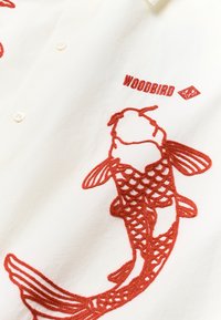 Chemise blanche avec un poisson koi rouge brodé et le texte "WOODBIRD" près du col, montrant les boutons et les motifs de couture détaillés.