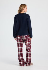 Vrouw die met haar rug naar de camera staat, gekleed in een marineblauw sweater, rode en witte geruite broek en beige slippers op een effen witte achtergrond.