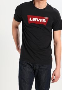 Černé bavlněné tričko s velkým červeným obdélníkovým logem Levi's uprostřed. Tričko má kulatý výstřih a krátké rukávy.