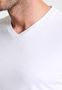Man’s lower face and neck with stubble wearing a plain white V-neck T-shirt against a white background.
