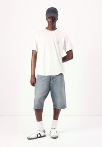 Tricou alb din bumbac cu un grafic mic roșu, asociat cu șorturi din denim albastru, șosete albe și pantofi sport alb-negru. Se poartă o șapcă gri.