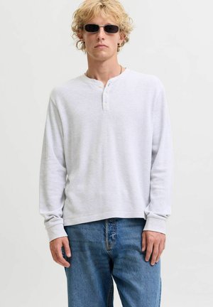 Personne blonde portant des lunettes de soleil rectangulaires noires, une chemise henley blanche à manches longues et un jean bleu, debout devant un fond clair uni.