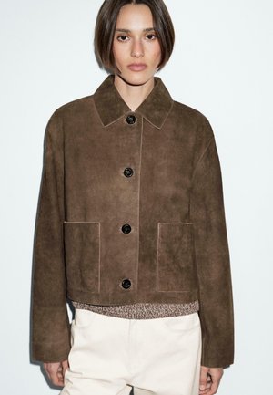 A model wears a brown suede jacket with large buttons, paired with a textured sweater and light pants, against a plain white background.