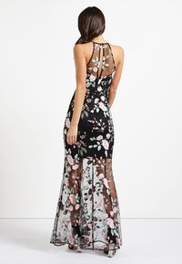 Robe noire à fleurs avec un superposition transparente, ornée de fleurs brodées roses et vertes. Design sans manches, dos nu, silhouette ajustée, longueur au sol.