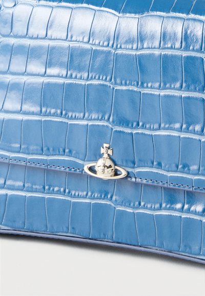 Surface en cuir embossé crocodile bleu avec un petit emblème argenté en forme de Saturne et un détail croix centré près du bord inférieur.