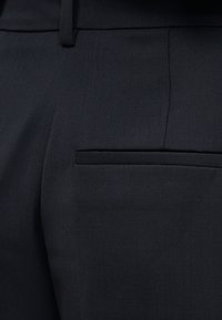 Κοντινό πλάνο μαύρου παντελονιού ραμμένου, που δείχνει την υφή του υφάσματος, τη θηλιά για τη ζώνη και την οριζόντια τσέπη στο πίσω μέρος.