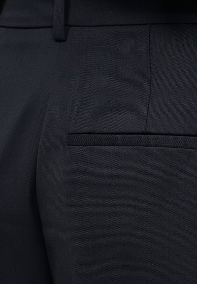 Zbliżenie na czarne, dopasowane spodnie pokazujące teksturę tkaniny, szlufkę na pasek oraz poziome tylne kieszenie.