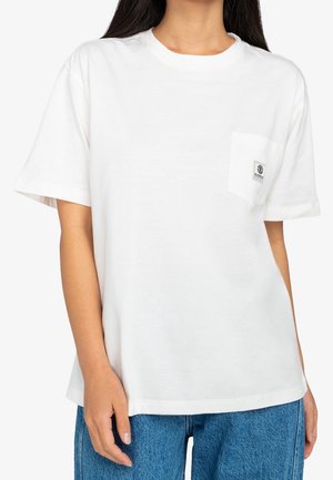 Femme portant un T-shirt blanc uni à manches courtes avec un petit logo sur la poche poitrine, associé à un jean bleu en denim.