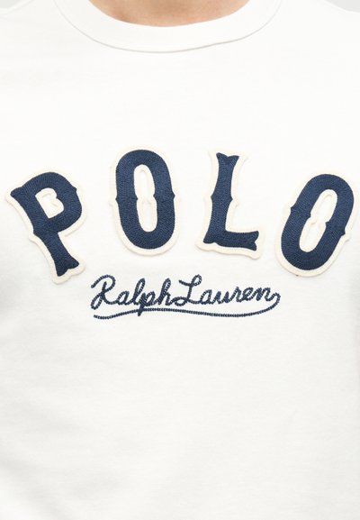 Λευκό βαμβακερό μπλουζάκι με κεντημένα γράμματα σε ναυτικό μπλε που γράφουν "POLO" και "Ralph Lauren" με καλλιγραφική γραφή από κάτω. Κοντά μανίκια και στρογγυλή λαιμόκοψη.