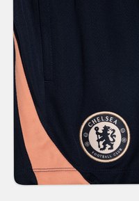 Tumesinised lühikesed püksid, millel on oranž külgpaneel, ning ümmargune brodeeritud märk, kus on lõvi kujutis ja tekst "CHELSEA JALGPALLIKLUBI".