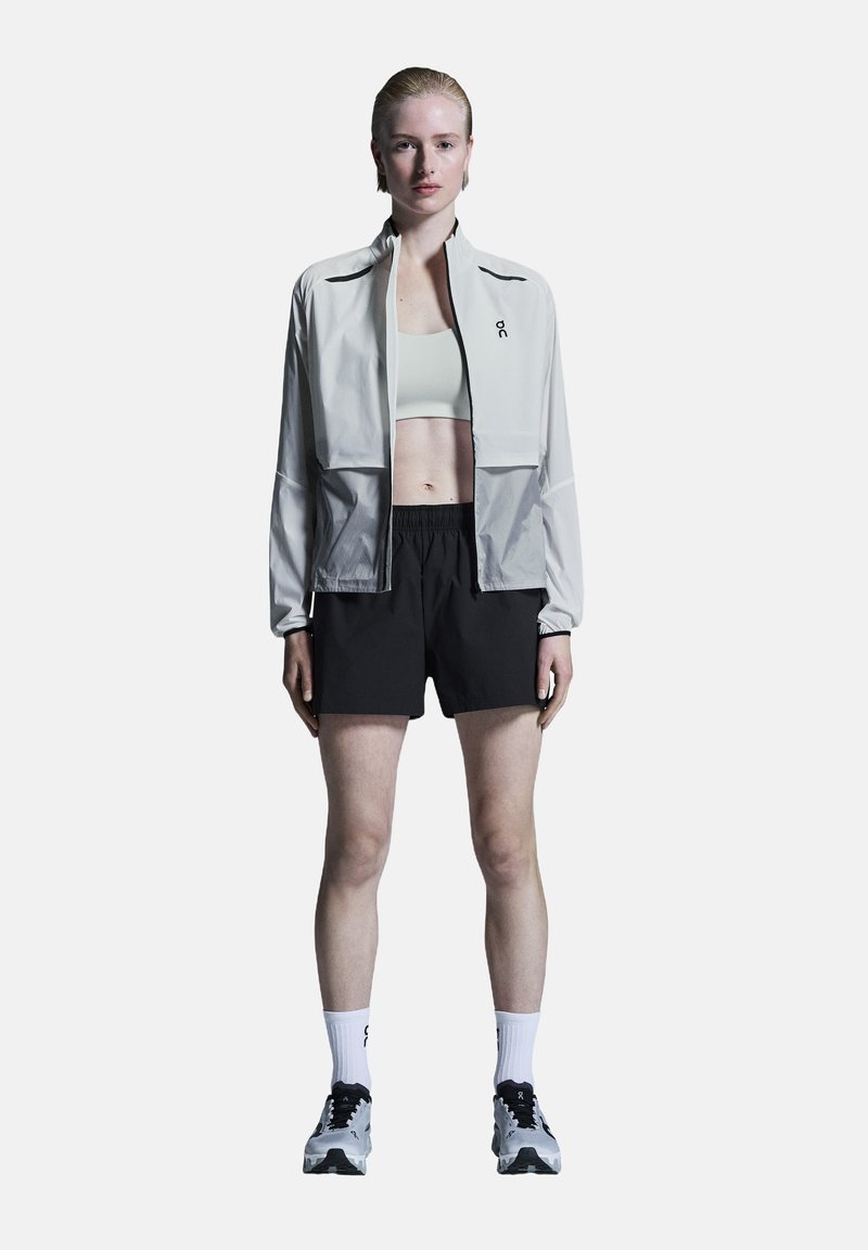 Veste zippée gris clair sur un crop top vert clair, associée à un short noir. Le mannequin porte également des chaussettes blanches et des chaussures de sport noires.