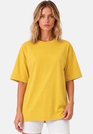 Vrouw met schouderlang blond haar die een oversized geel kortärmelig overhemd en een witte korte broek draagt, staand tegen een effen achtergrond.