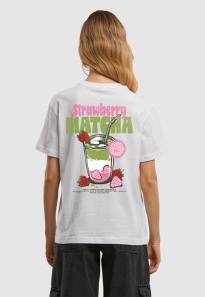 Person med langt blondt hår iført en hvid t-shirt med en grafik af en jordbær matcha-drink med jordbær og isterninger.