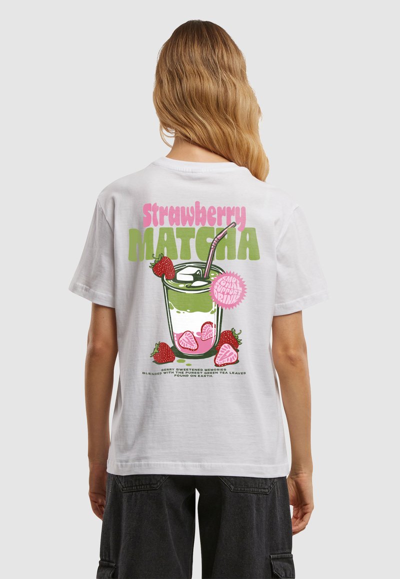 Personne aux longs cheveux blonds portant un t-shirt blanc affichant un graphique d'une boisson matcha à la fraise avec des fraises et des glaçons.