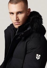 Czarna kurtka puchowa z kapturem wykończonym futrem, zapięciem na zamek i naszywką z logotypem na lewym rękawie. Teksturowany materiał, minimalistyczny design.