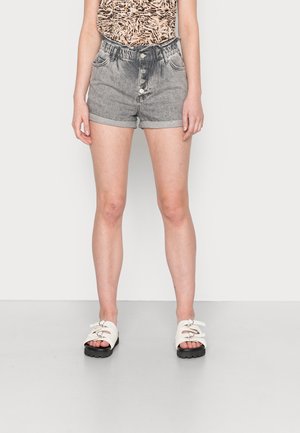 Shorts en denim avec un délavage gris clair, fermeture à cinq boutons à l'avant et ourlets roulés ; associés à des sandales blanches avec un grand matériel décoratif.